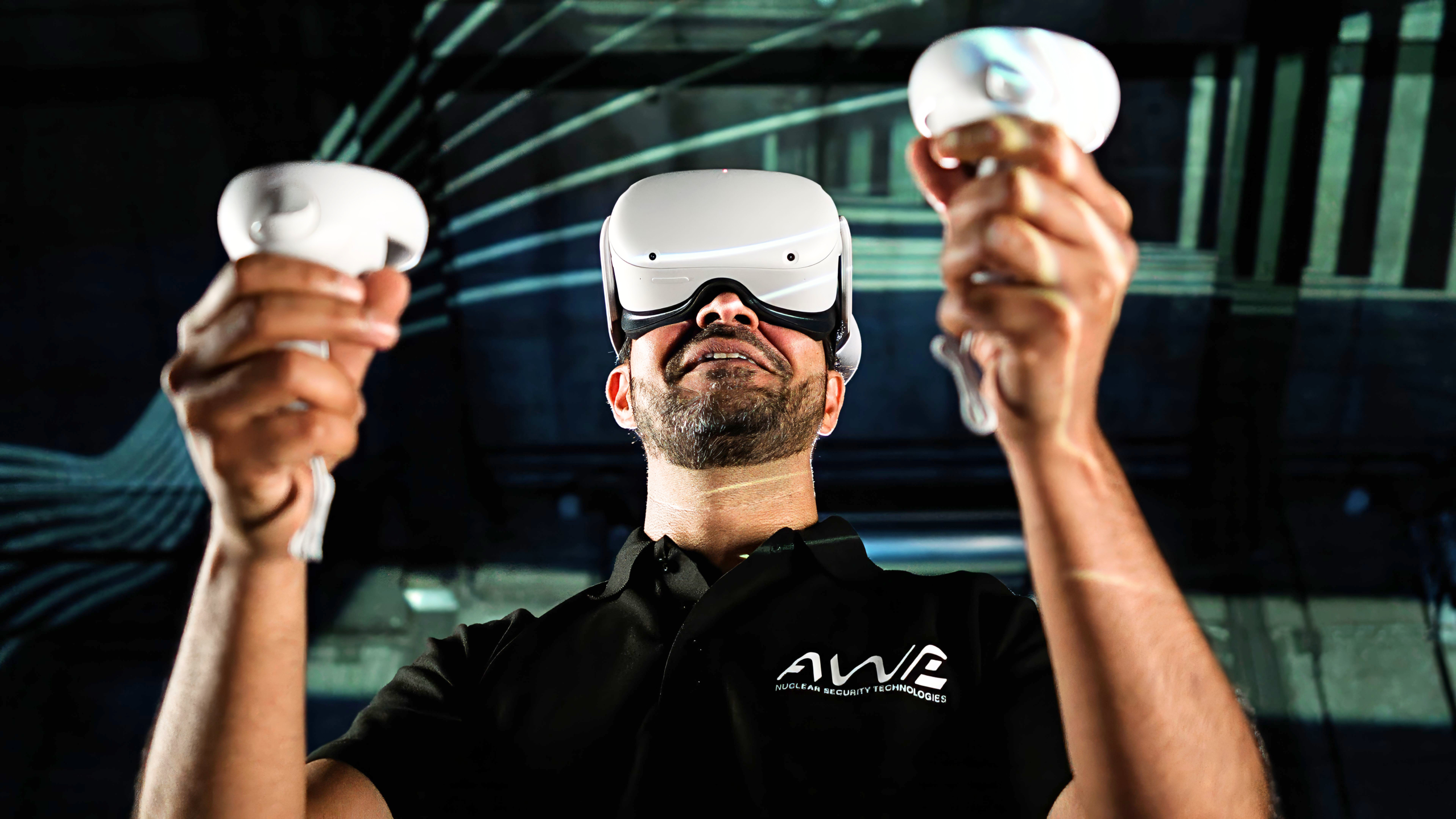 Person wearing a virtual reality headset and holding two VR controllers in an indoor setting featuring abstract lines and shapes; wearing a black 'AWE' shirt.