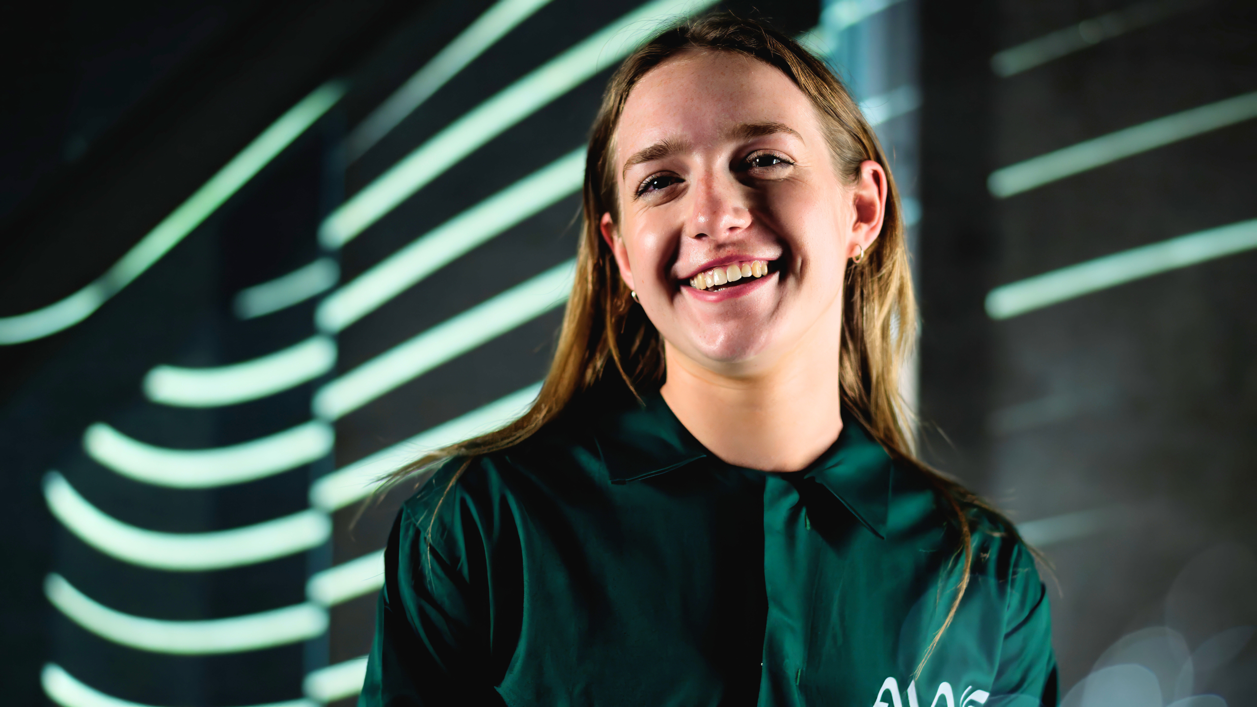 A woman in a dark green shirt with "AWE" on the chest stands in front of a dark background with curved, illuminated lines.