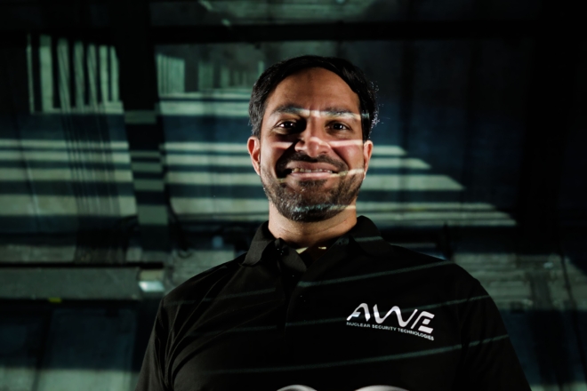 The image depicts a person standing in a dimly lit setting, with shadows and light patterns creating a moody background. The individual's face is intentionally blurred for privacy. They are wearing a black polo shirt featuring the logo