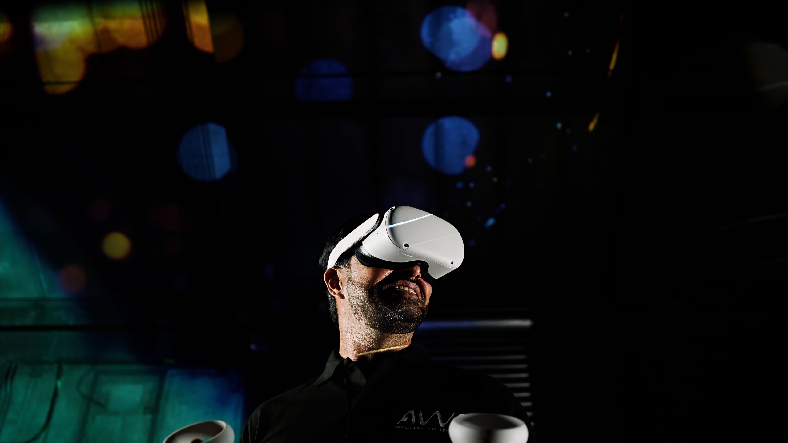 The image features a person wearing a VR headset and a dark shirt with the letters "AWE" visible on the chest. The background is dark and features a bokeh effect with colourful, out-of-focus lights, giving the image a visually striking and atmospheric quality.