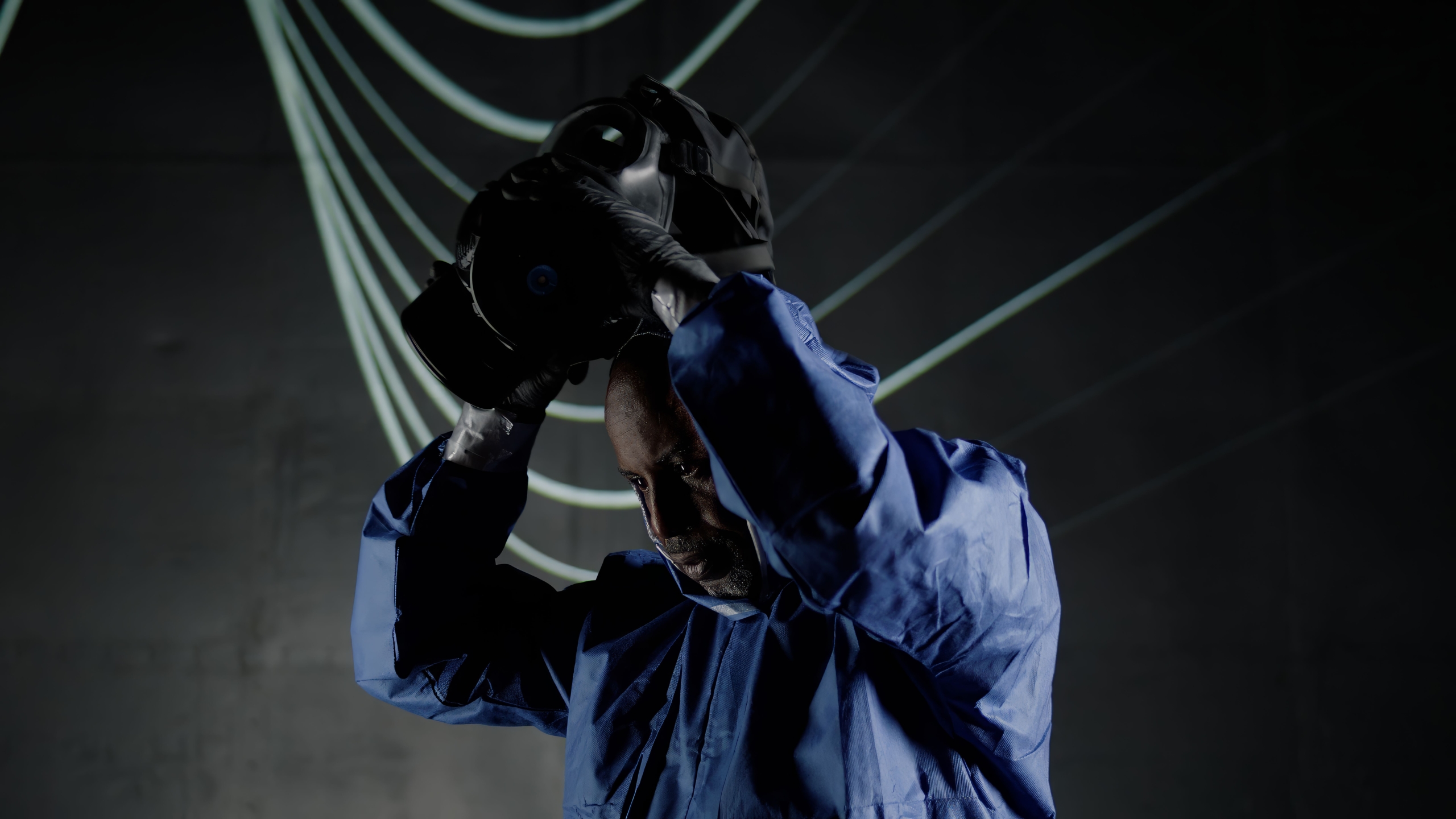 Person in a blue protective suit and black gloves holding a virtual reality headset above their head, with curved illuminated lines in the dark background.