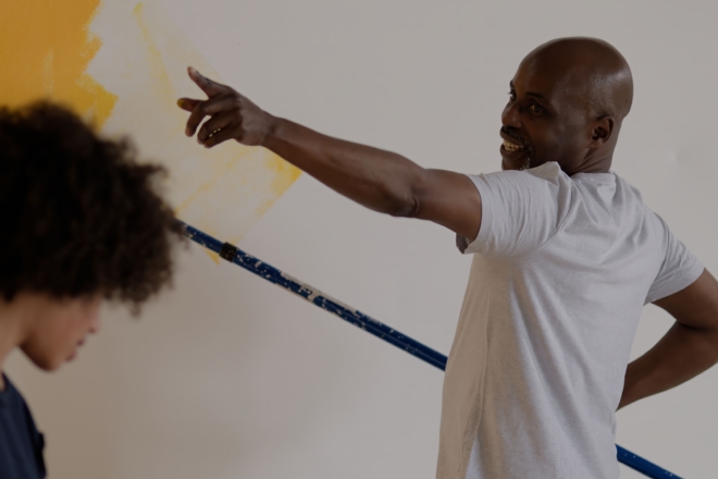 Two people painting a wall; one holds a roller with yellow paint and points toward the wall, while the other looks down. The wall is partially painted yellow.