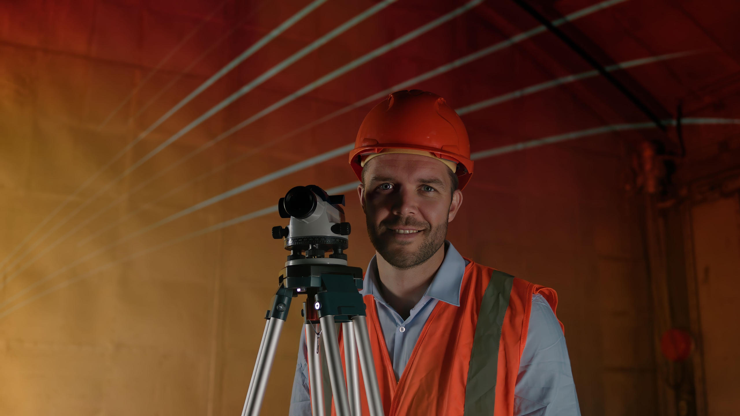 A person in an orange hard hat and safety vest smiles at the camera and uses a surveying instrument on a tripod in a dimly lit industrial setting