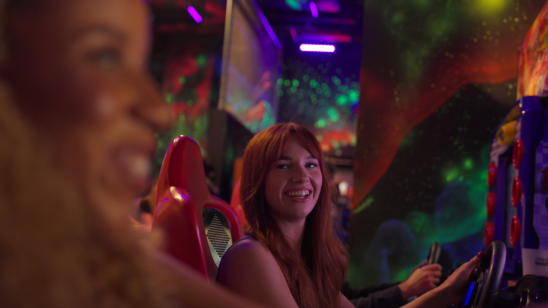 A group of people playing racing games in an arcade, seated in gaming chairs with steering wheels, surrounded by vibrant neon lights and a cosmic-themed mural.
