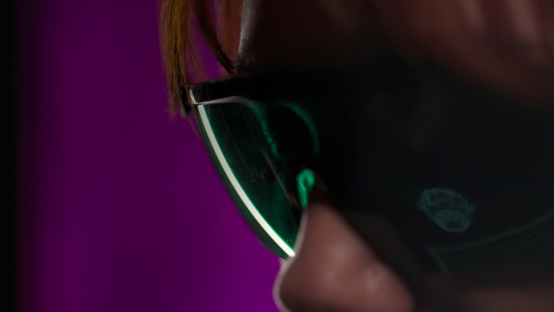 Close-up of a woman wearing dark sunglasses with green-tinted lenses, set against a solid purple background. The focus is on the sunglasses and their reflection.