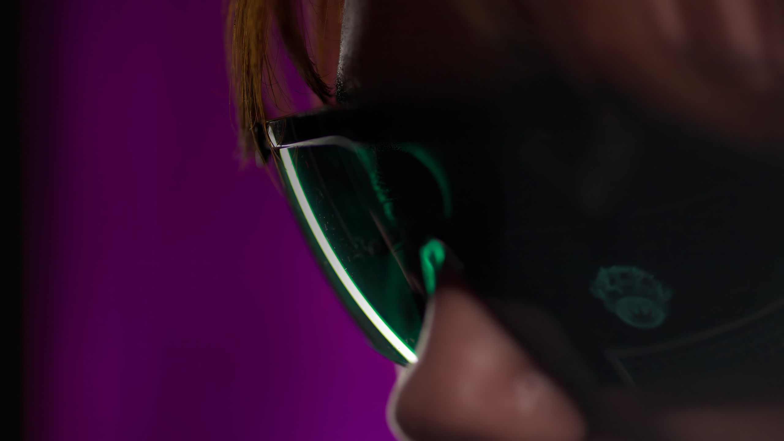 Close-up of a woman wearing dark sunglasses with green-tinted lenses, set against a solid purple background. The focus is on the sunglasses and their reflection.