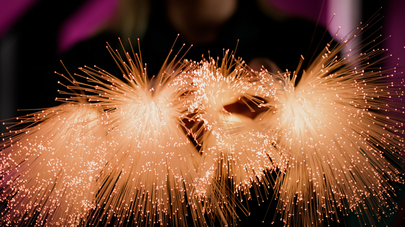 A person holds glowing bundles of fibre optic cables, with illuminated ends creating a bright, colourful light display against a dark background.