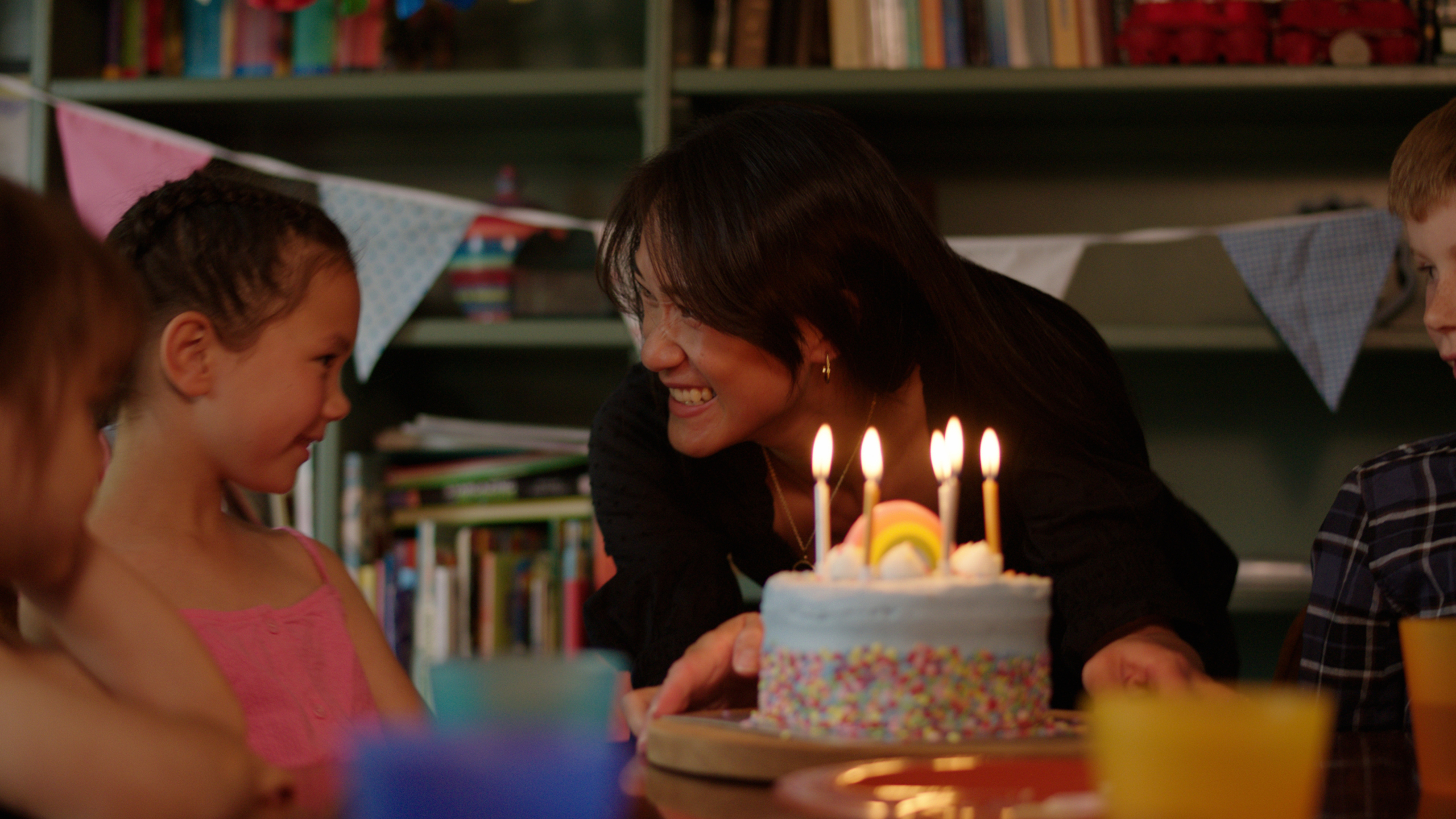 A woman smiles at a child where all are gathered around a table with a birthday cake topped with five lit candles, surrounded by bookshelves and colourful bunting.