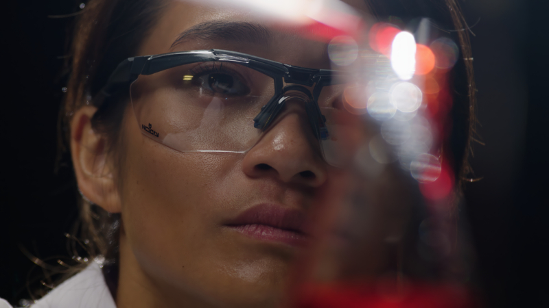 A scientist with goggles looks at a blurry flask with soft lights in the background.