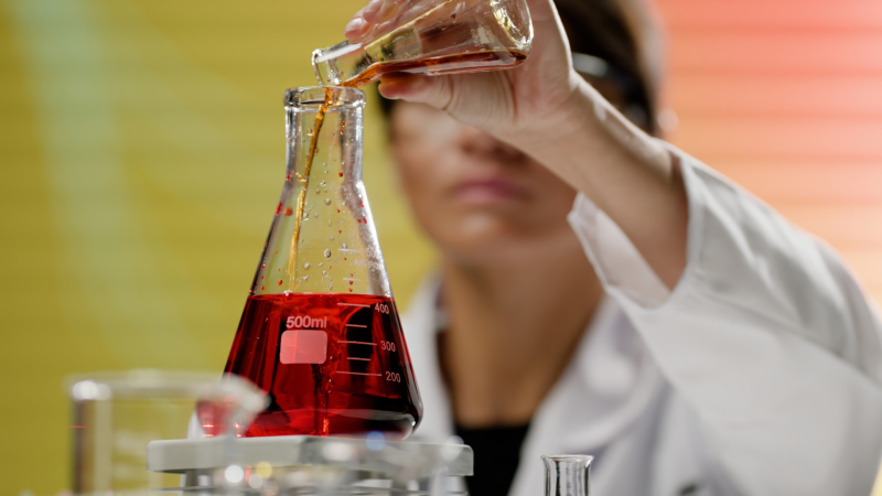 A woman in a lab coat pours liquid into a 500 ml Erlenmeyer flask with red solution, set against a blurred yellow-orange background.
