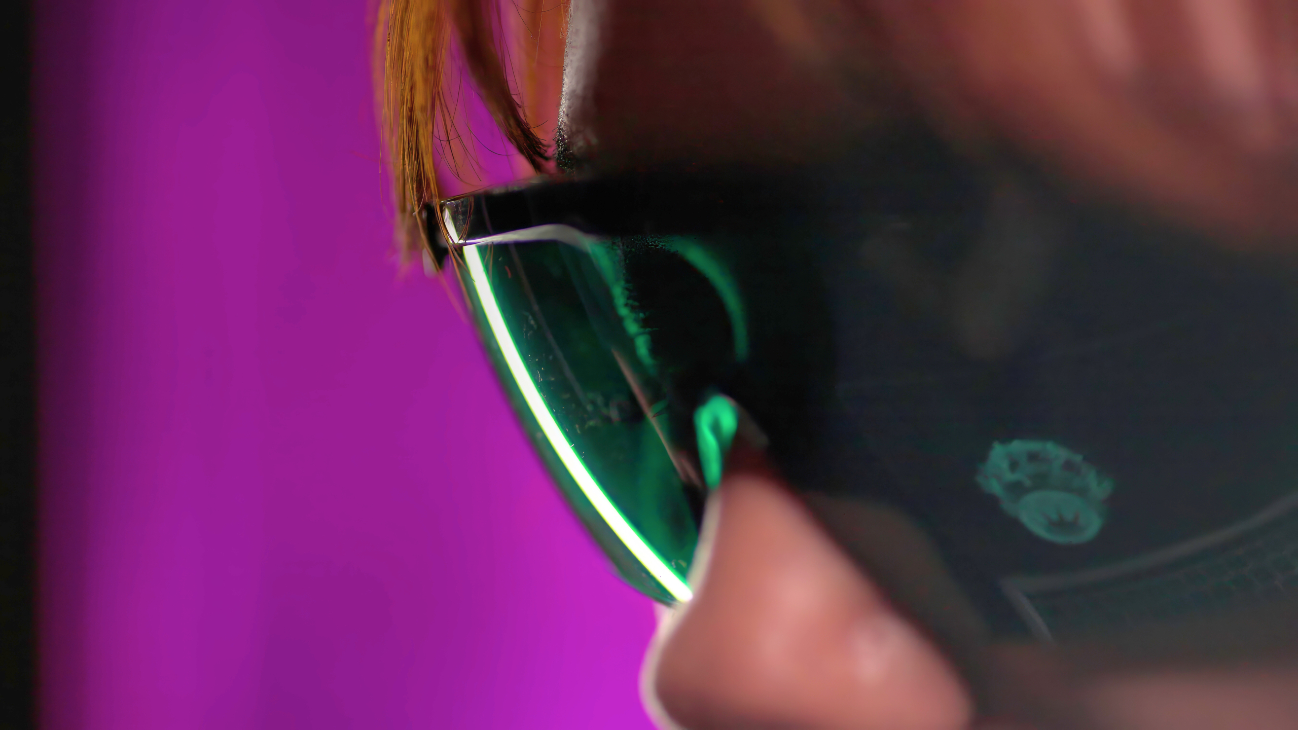 Close-up of a woman wearing dark sunglasses with green-tinted lenses, set against a solid purple background. The focus is on the sunglasses and their reflection.
