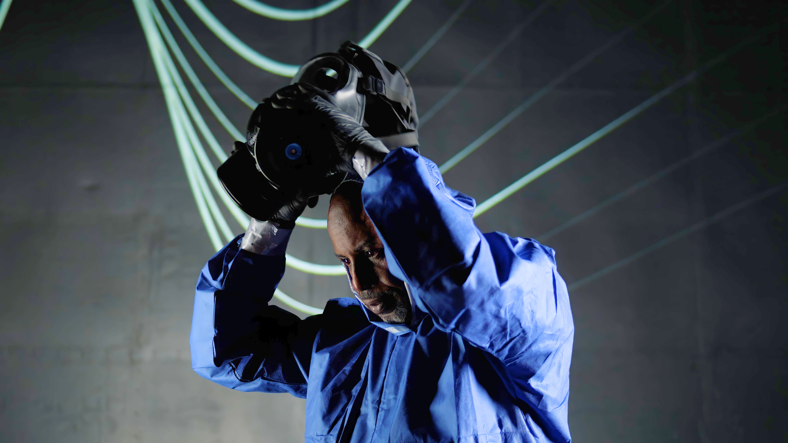 Person in a blue protective suit and black gloves holding a virtual reality headset above their head, with curved illuminated lines in the dark background.