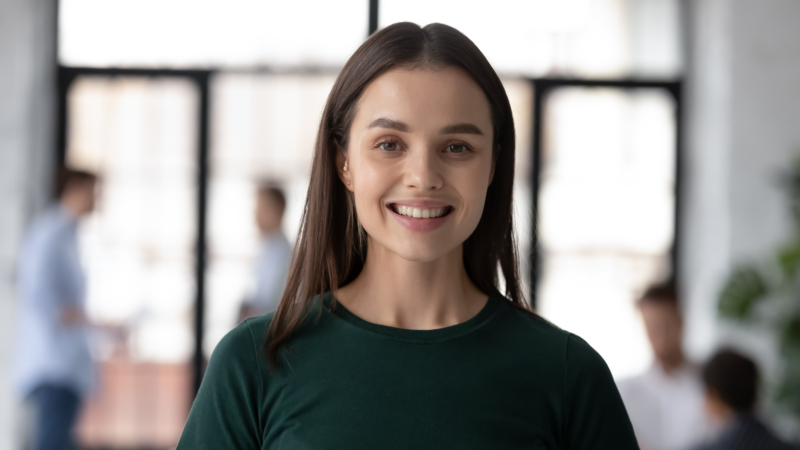 A person with long, straight hair wearing a dark green shirt stands indoors. The background is blurred, showing other people in a room with large windows
