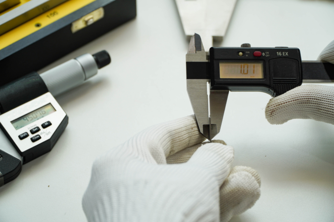 Person wearing white gloves using a digital caliper to measure an object, with the display reading '10.71'; additional measuring tools like a micrometer and spirit level are visible in the background.