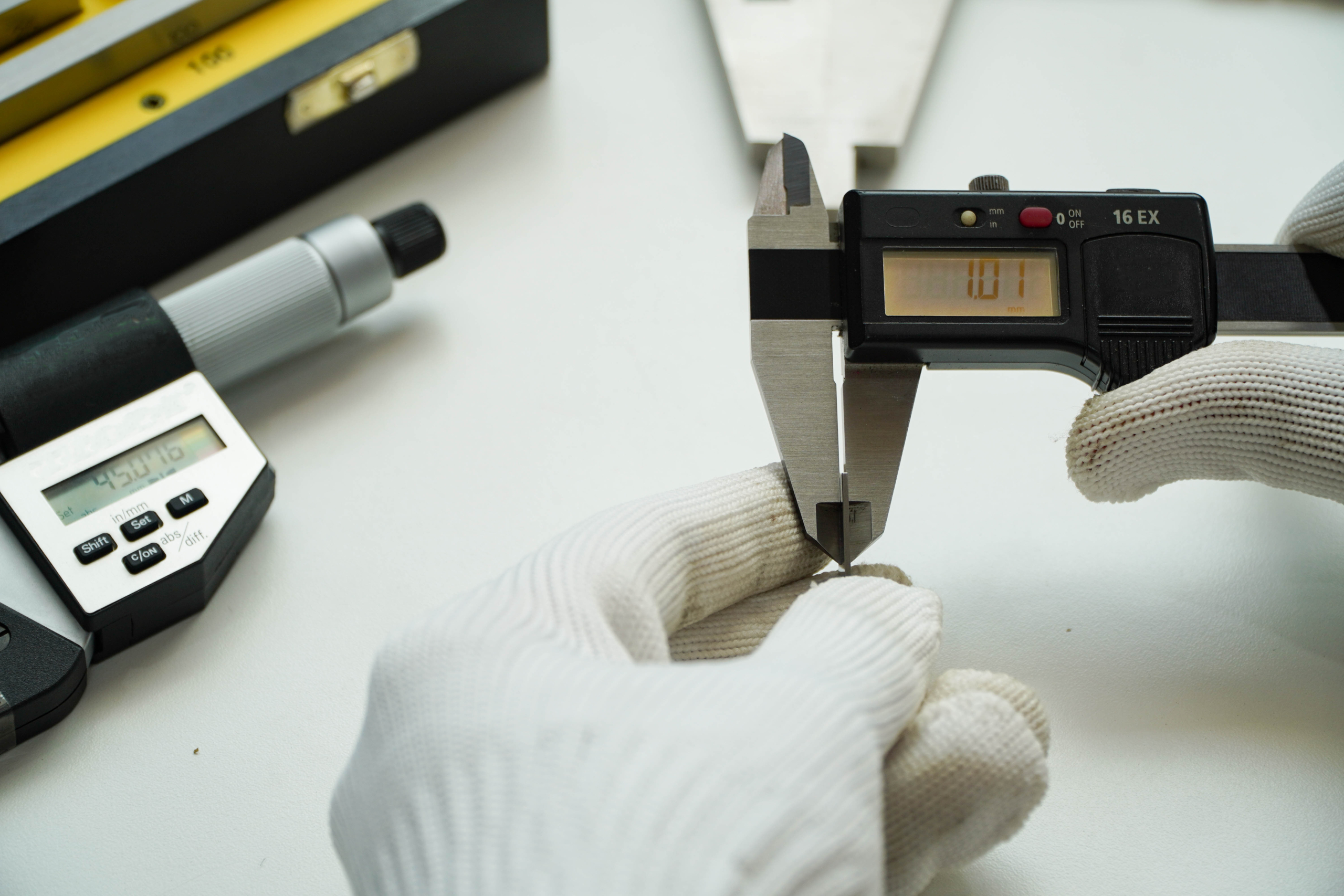 Person wearing white gloves using a digital caliper to measure an object, with the display reading '10.71'; additional measuring tools like a micrometer and spirit level are visible in the background.