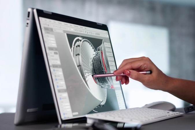 A person using a stylus to interact with a 3D model of a jet engine on a convertible laptop in tent mode, with detailed engine components visible on the screen and a white keyboard in front.