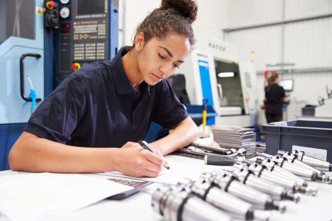 A person in an industrial setting sits at a table writing on papers, with several metal cylindrical components in front of them. Machinery and another worker are visible in the background.