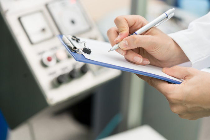A person holding a blue clipboard and writing with a pen, standing in front of a control panel with various buttons and dials.