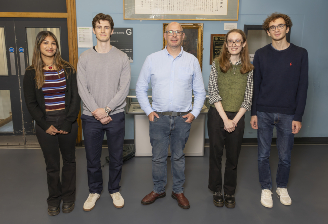 Five people standing indoors in front of a blue wall with framed documents and plaques. They are dressed casually and facing the camera, standing on a grey floor near a door with glass panels.