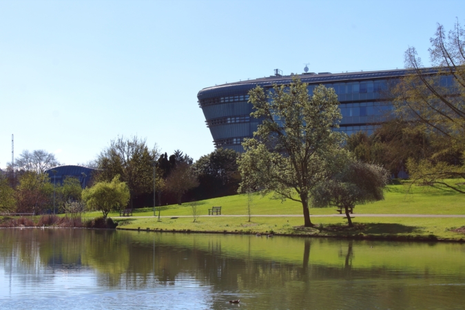 Curved glass building on a grassy hill with trees, reflected in a pond under a clear blue sky.