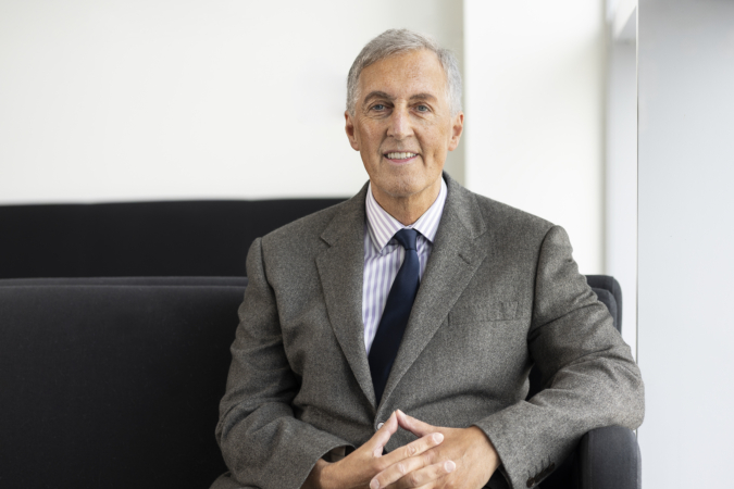 Scott McCulloch wearing a grey suit jacket, light blue striped dress shirt, and dark tie, seated on a dark sofa with hands clasped, in a bright modern office setting