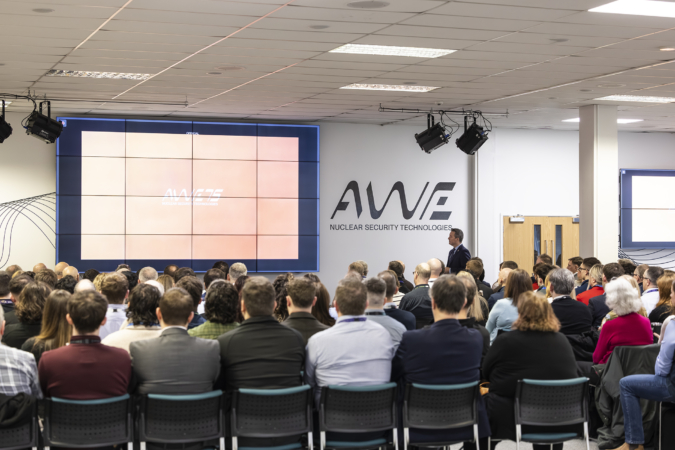 Feature image Group of people sat down facing away from the camera sat in rows at a conference watching a stage with a large screen