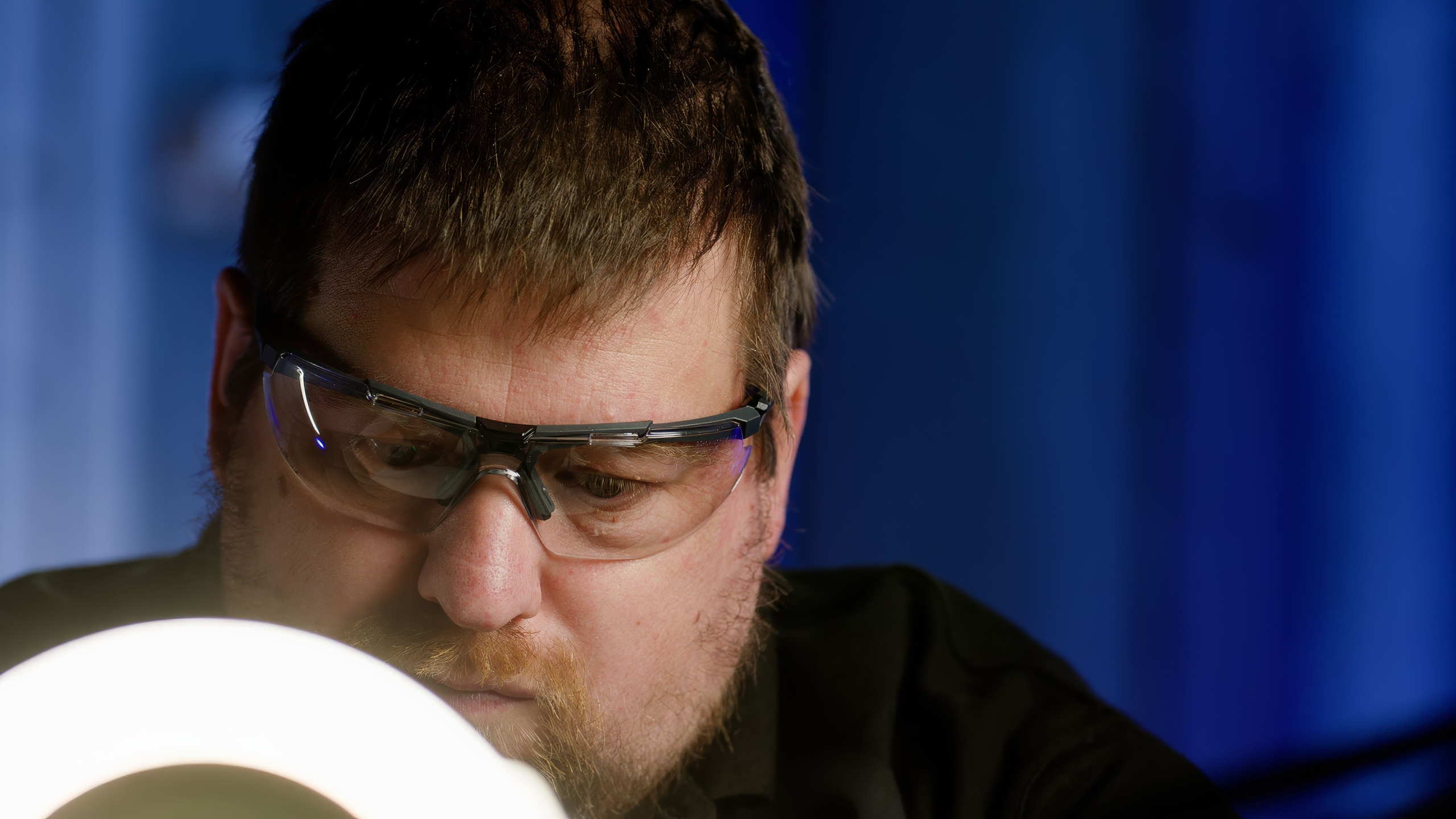 A person seated indoors with a dark shirt, illuminated by a circular light source in the foreground. The background features a deep blue curtain or wall, creating a professional and focused setting.