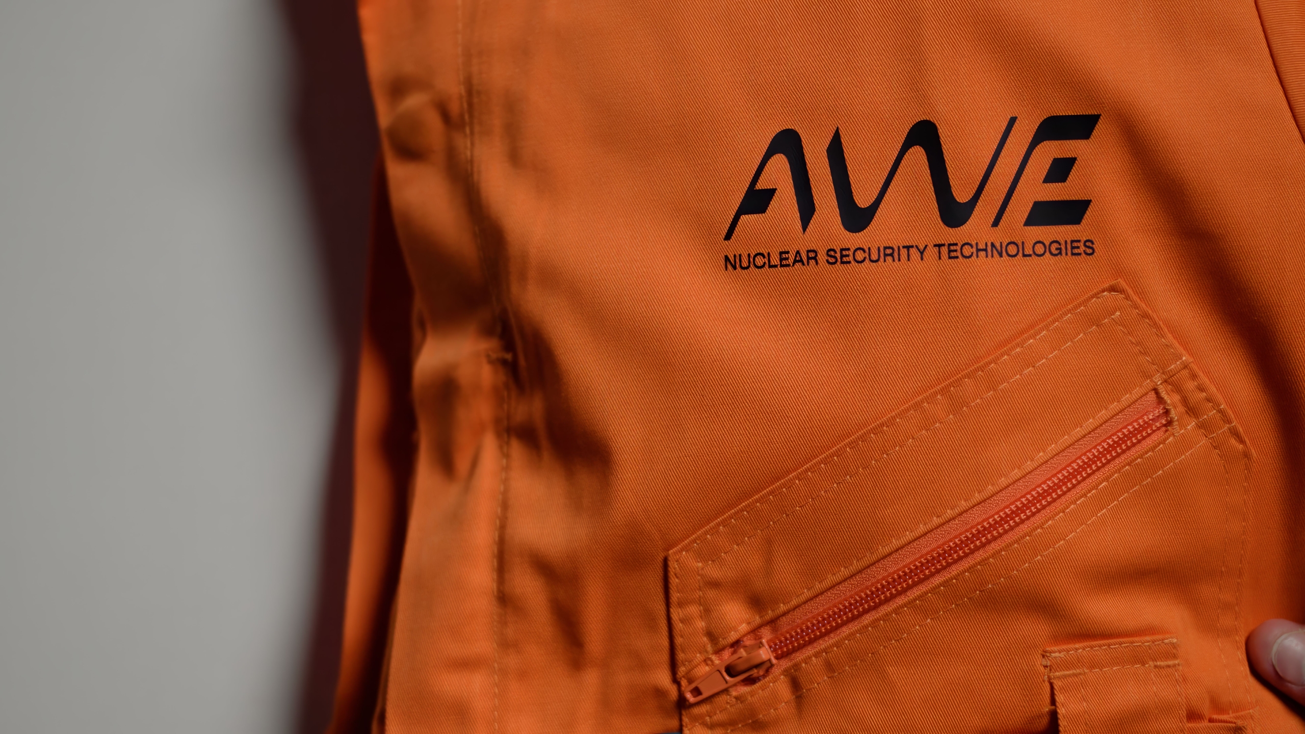 Close-up of an orange protective suit featuring the AWE logo and the words “Nuclear Security Technologies.” A zippered pocket is visible on the front, highlighting the branded workwear used in high-security engineering environments.