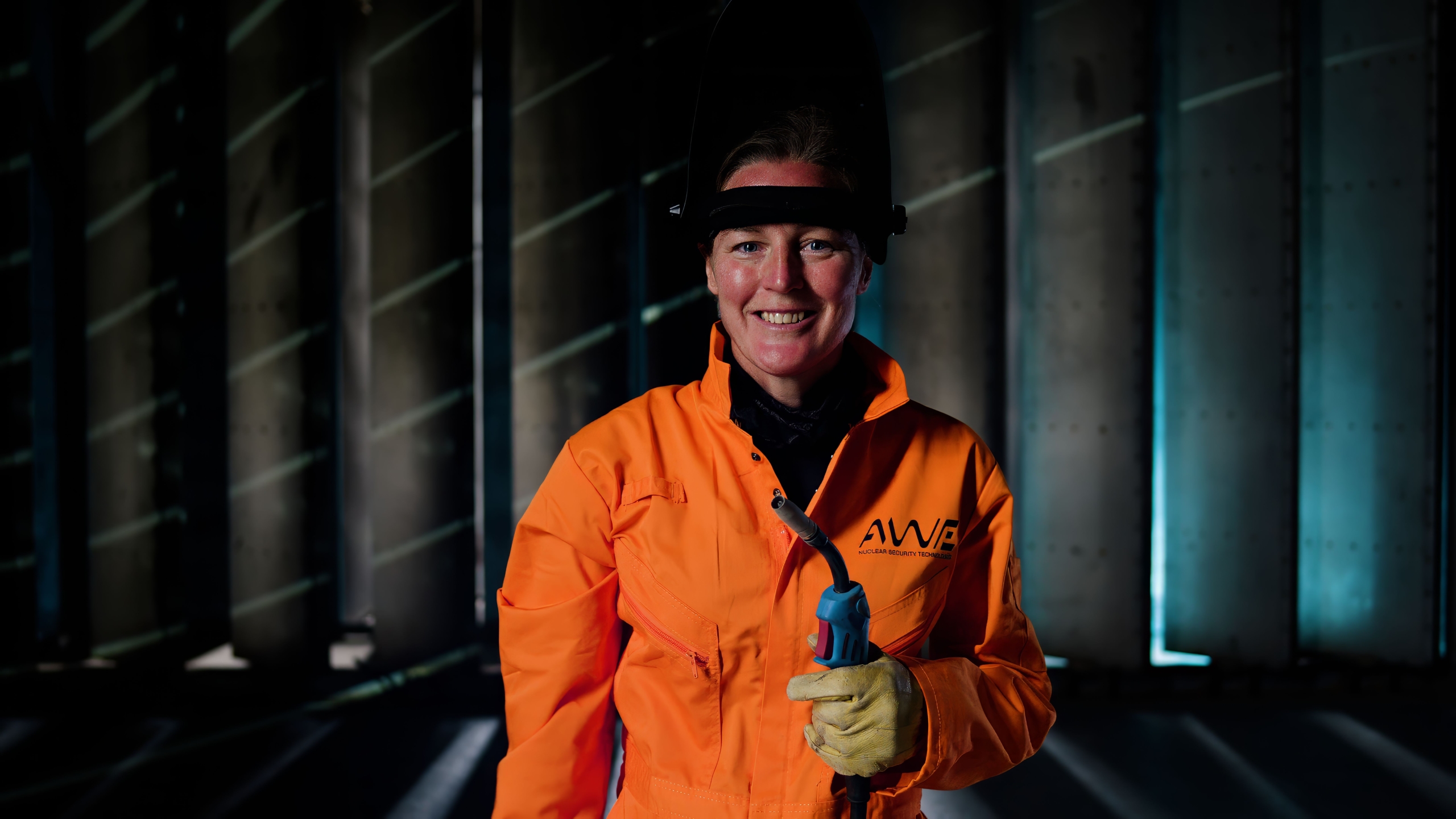 Person wearing an orange protective suit with AWE branding, holding a welding torch in a dimly lit industrial environment. Vertical metal panels and blue-toned lighting create a structured, high-security setting.