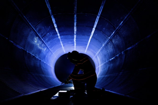 Silhouette of a person working inside a large cylindrical tunnel, illuminated by blue light beams converging toward the center. The figure is kneeling and engaged in a technical task, creating a dramatic industrial scene.